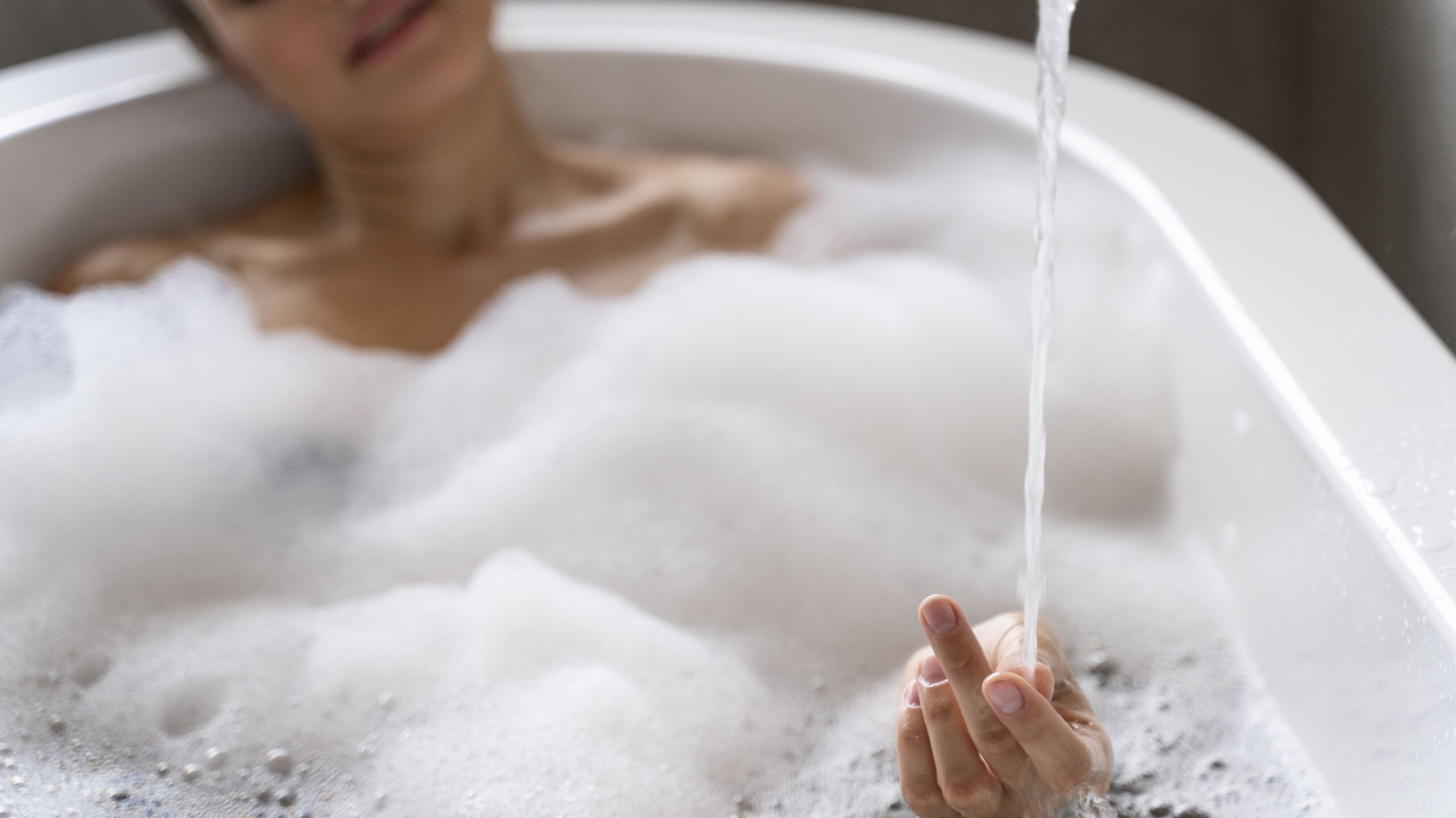 Person relaxing in a bathtub filled with bubbles, with water running and soft natural light in a quiet bathroom